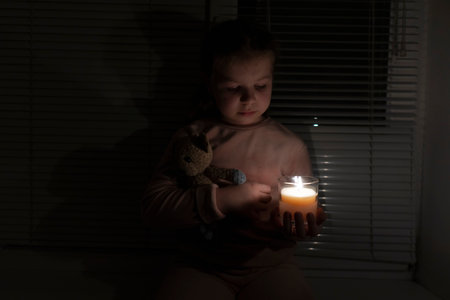 A girl in a dark room near the window holds a candle and a teddy bear in her hands. power outage concept. blackout. energy crisis. Destruction of infrastructureの写真素材