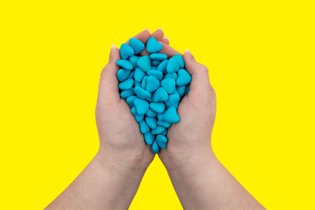 A handful of blue heart-shaped candies in hands isolated on a yellow background. Close-up, top view