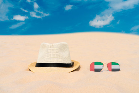 Sunglasses with glasses in form of flag of United Arab Emirates and a hat lie on sand against blue sky. Concept of summer holidays, travel and tourism in United Arab Emiratesの写真素材