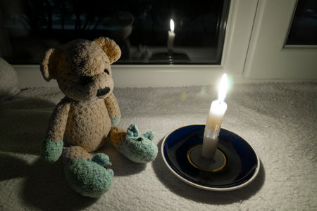 Blackout. A teddy bear sits in the dark on the windowsill near the window next to a burning candle. Power outputの写真素材
