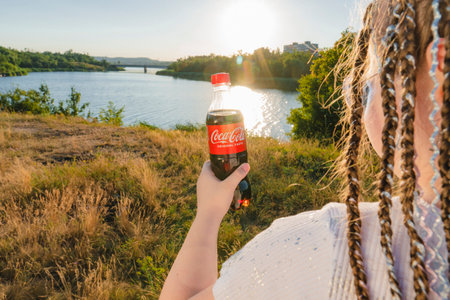 Krivoy Rog, Ukraine, June 26, 2024: A girl stands on the bank of the river and holds a bottle of Coca-Cola in her hand (view from the back). Summer sunny day. Outdoor recreationのeditorial素材