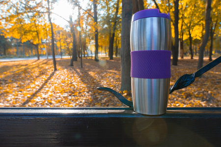 A stainless steel thermos mug with a purple band sits on a park bench, surrounded by vibrant golden...