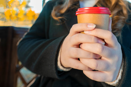 A cozy moment on a sunny autumn day with hands holding a warm paper coffee cup with a bright red lid...