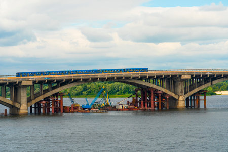 A train with blue and yellow cars moves along a large concrete bridge over a wide river on a cloudy summer dayの写真素材