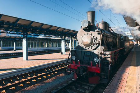 A vintage black steam locomotive with a red bumper stands on a railway track at a station under a blue skyの写真素材