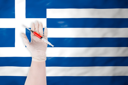 A hand in a protective glove holding a syringe with red liquid against the waving flag of Greece, symbolizing vaccination and medicineの写真素材