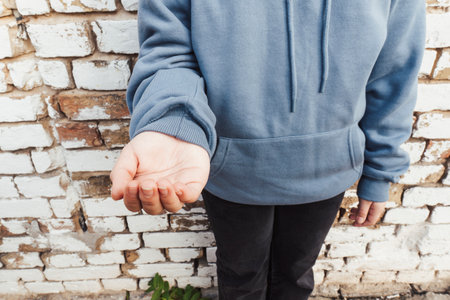 Sad homeless child in blue hoodie holding out an empty hand while standing near a weathered white brick wall. Concept of poverty and social crisisの写真素材