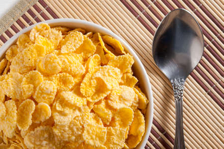 A white bowl of cornflakes, next to a metal spoon. Everything is on a napkin.