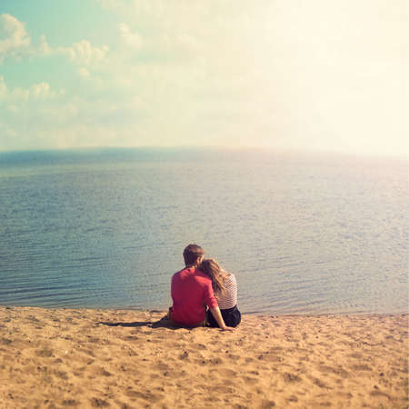 young couple sitting embracing on the sand looking out to seaの写真素材