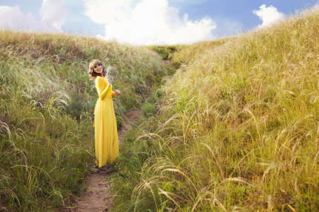 Outdoor portrait of a beautiful brown hair woman. Attractive girl on the feather grass meadow with blue cloudy sky in yellow dress. Warm colors landscape horizontal photoの写真素材