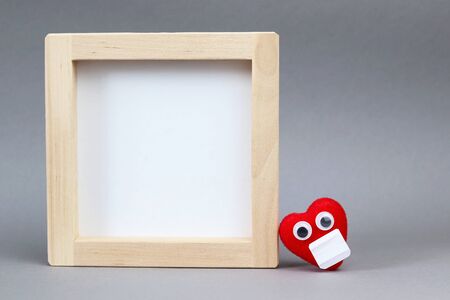 Wooden frame for text and a red heart in a mask on a gray background. Copy space - hygiene concept, health, quarantine, coronavirus, new normal lifestyleの写真素材