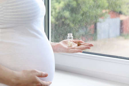 A pregnant, sick second-trimester woman in a white T-shirt is holding a glass jar of pills near a...