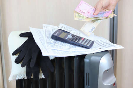 Hryvnia in hand. Defocused bills and calculator, scarf and gloves on a battery. Copy space - the concept of paying for utilities, heating, cold, money, savings, homeの写真素材