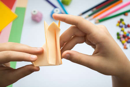 Children's hands do origami  wolf  from peach paper. Working place is decorated with leaves of color paper, scissors, colored wood ladybugs and pencils.の写真素材