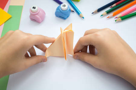 Children's hands do origami  wolf  from peach paper. Working place is decorated with leaves of color paper, scissors, colored wood ladybugs and pencils.の写真素材