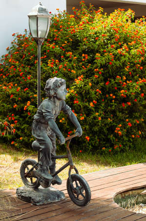 The outside lapm with the statue of a boy riding a bicycle as a exterior detail of a garden over the background of bush with orange flowersの写真素材