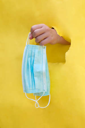 the vertical photo of the hand throught the hole in the yellow background offering the blue disposable masksの写真素材
