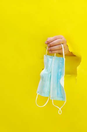 vertical photo of a young hand giving the blue disposable masks through a hole in the yellow backgroundの写真素材