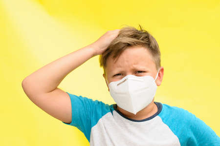 Horizontal portrait of an unhappy boy with the disposable mask on the face holding the head with one hand over the yellow backgroundの写真素材