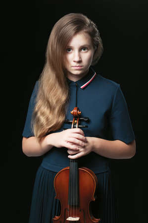 A teenage caucasian girl in a dark blue dress holding a violin infront of her body over the dark backgroundの写真素材