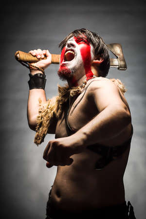 Portrait of man in war paint, with an ax in his hand, a warrior brandishing an ax up furiously screaming, barbarian of ancient times, Viking and savage.の写真素材
