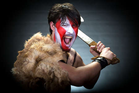 Portrait of man in war paint, with an ax in his hands, a warrior brandishing an ax furiously screaming, barbarian of ancient times, Viking and savage.の写真素材