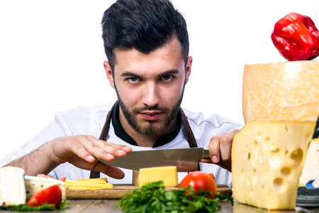 Young chef model looks, cooks of the beneficial ingredients. The man in an apron cuts the cheese into thin slices  looking into the cameraの写真素材