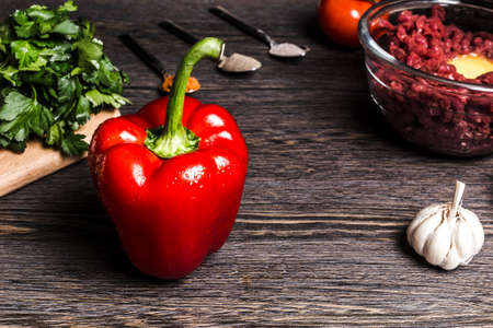 pepper paprika on a wooden background near parsley, garlic, minced meat, tomato and spoons with spices, the concept of cooking in the kitchenの写真素材