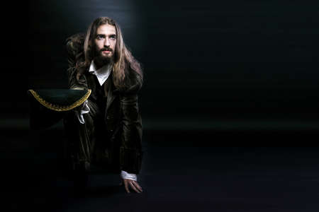 studio photography medieval wary pirate man with long hair and beard, sat down on one knee looking...