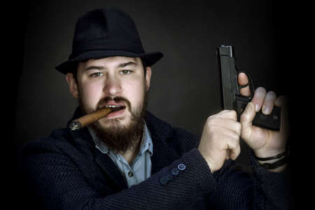 bearded gangster in a hat with a gun in his hand and a cigarette in his mouth on black background