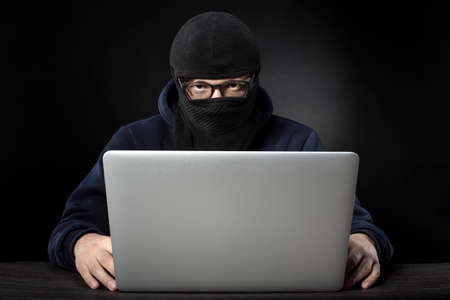 terrorist in mask and glasses sitting at a laptop on a dark backgroundの写真素材
