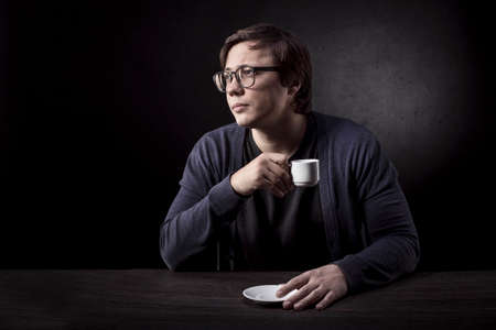 man in glasses drinks coffee and thoughtfully looks away on the background a dark concrete wall
