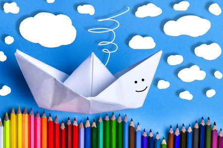 a boat of paper on a blue background near to colored pencils, concept of swimming on the wavesの写真素材