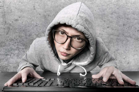 a hacker (programmer) in a hood and glasses against a concrete wall looks strangely at the cameraの写真素材