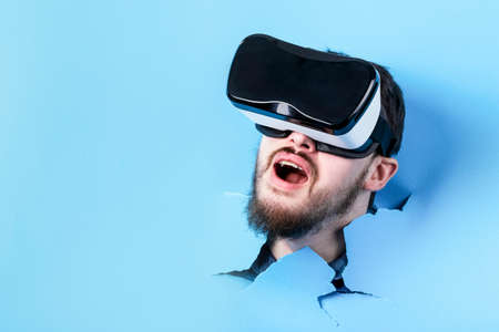 Bearded man making hole in blue paper. Surprised man in virtual reality glasses. VR device.