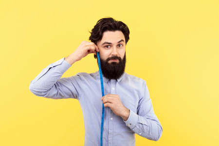 studio portrait of a bearded hipster man measures the length of the beard with measuring tape, over yellow backgroundの写真素材