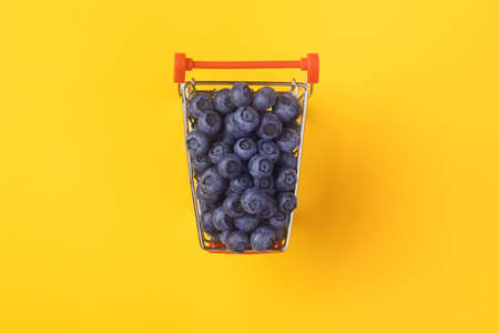 Shopping cart filled with blueberries on yellow background. Healthy shopping and eating concept.