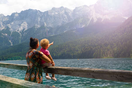 Young kid girl with mom sitting by the lake side admiring Alps, little girl pointing a finger to the top of the mountain, concept of traveling and conquering new countriesの写真素材