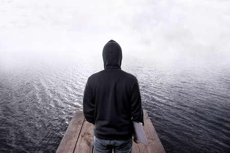 back view of hacker in the hood, with laptop in the hand, on wooden pier going into the water and...