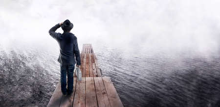 Back view of a man in black hat, holding business case, standing on wooden pier and looking into the...