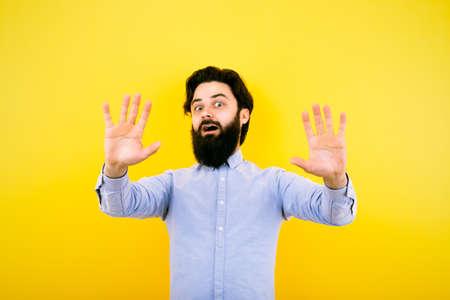 Portrait of shocked bearded man keeps hands up. Surprised guy in casual shirt on yellow background....