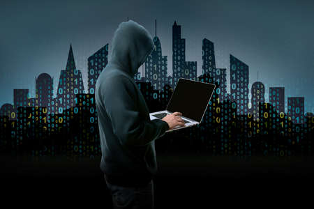unknown hacker in the hood with laptop over background of city silhouette with binary code