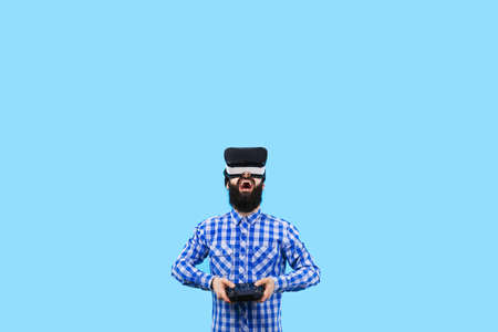 surprised bearded man in VR (virtual reality glasses) with remote control in hand controls the drone, image on blue backgroundの写真素材