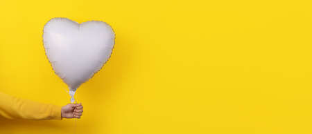 white foil balloon in shape of heart in hand over yellow background, panoramic layoutの写真素材