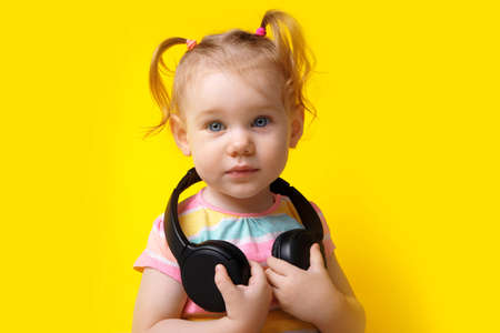 cute caucasian baby with wireless headphones, little girl looking at the camera over yellow...