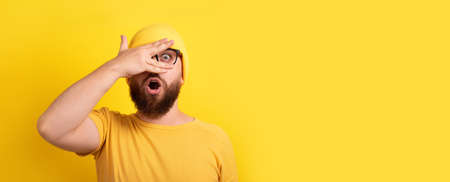 shocked man looking through fingers over yellow background, panoramic layout with space for textの写真素材