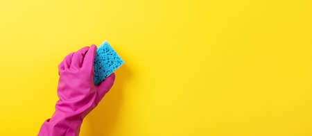 A hand in a pink rubber glove holds a blue washing sponge over yellow background, panoramic layoutの写真素材