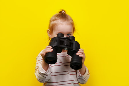 little girl looking into binoculars over yellow backgroundの写真素材