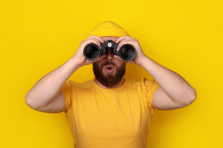 surprised man looking into binoculars over yellow backgroundの写真素材