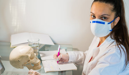 young female doctor or medical student holding human skull, pen and reading anatomy and writing on textbookの写真素材
